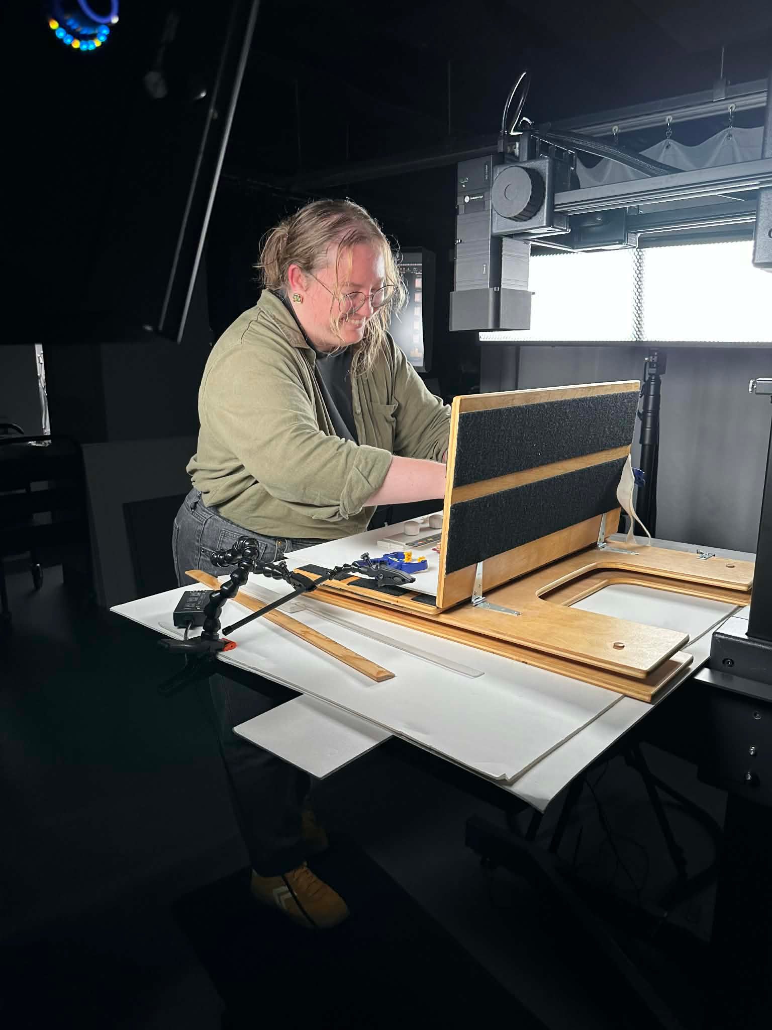 A photograph of a woman adjusting a support holding a manuscript book. Bright light shines down on the setup and black curtains surround them.
