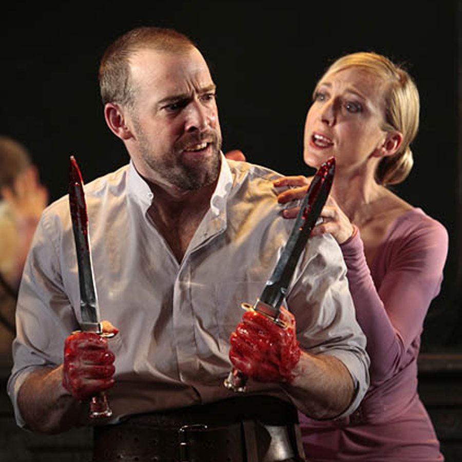 A man holding two bloody knives with a woman touching his shoulder