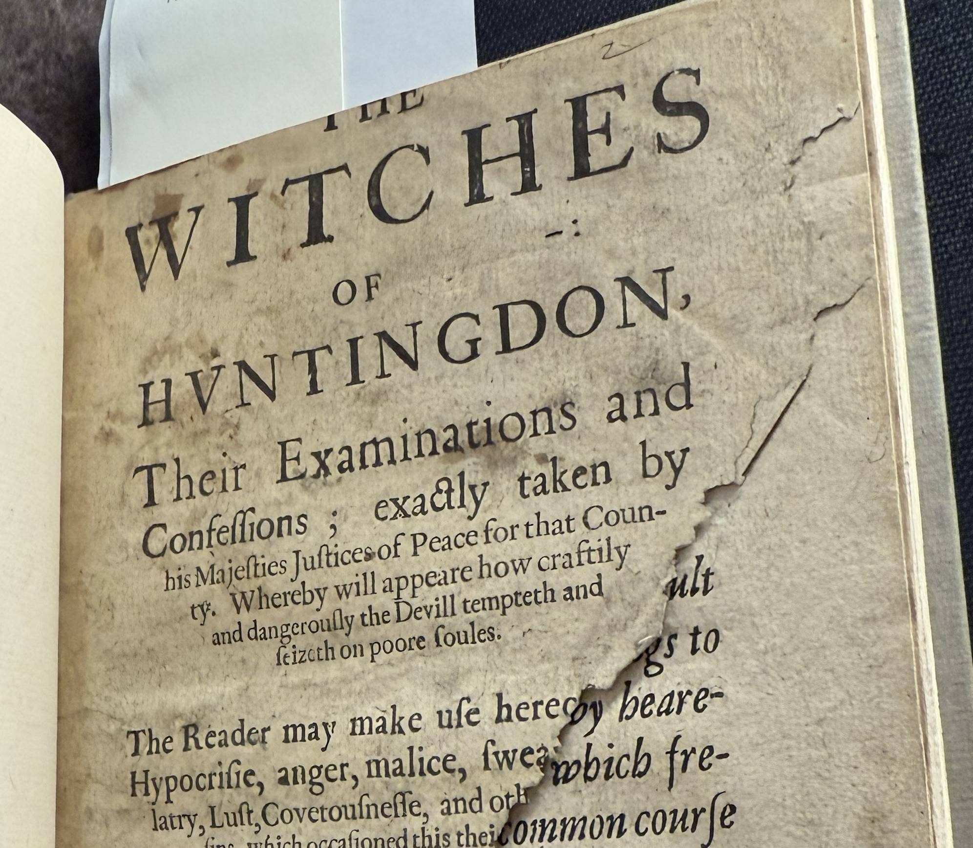 A printed title page of a book that is missing a large triangle shaped part of the bottom right of the page, revealing the text page of printed text underneath.