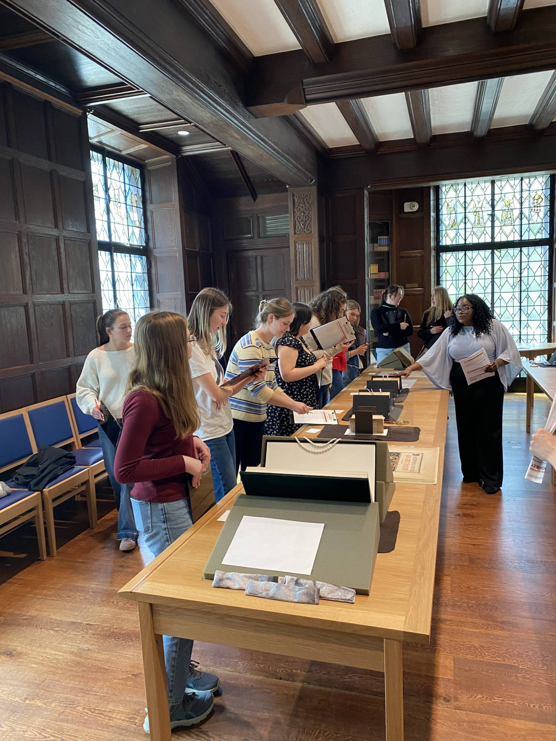 Students with clipboards in hand lean over books in cradles on a table while a instructor walks by, pointing at one of the items