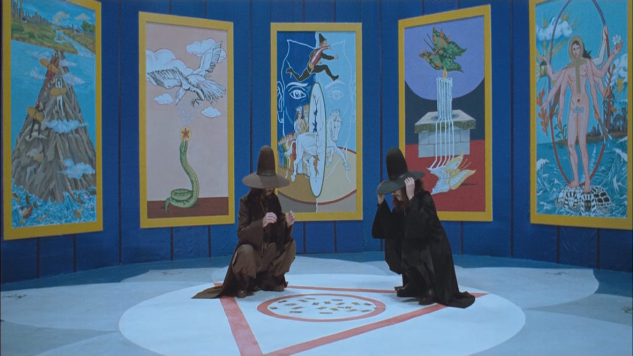 Two men in robes and tall round hats squat in a triangular figure on the floor. Surrounding the men on the blue walls of the room are five paintings with symbolic imagery.