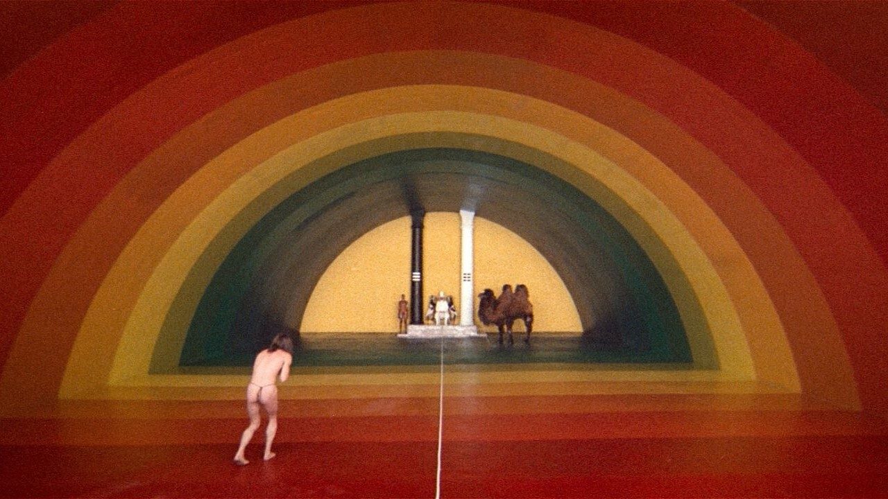 A film still showing a mostly naked man walking towards a rainbow-tunnel at the end fo which waits a man seated between black and white pillars, a camel on one side and a human-figure on the other.