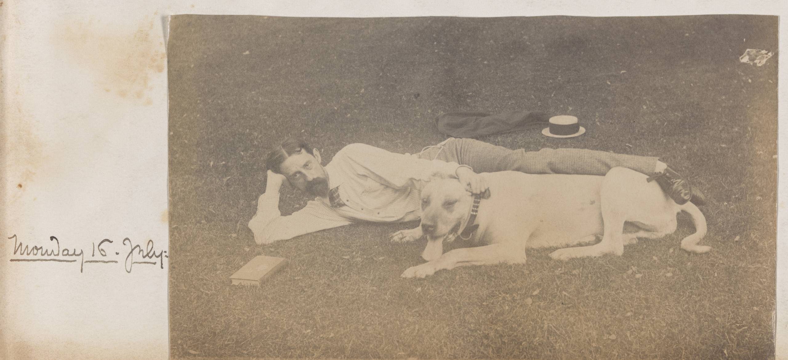 A faded sepia-toned photograph of a bearded man stretched out beside a big tan or white dog. The photograph is captioned Monday 16. July: