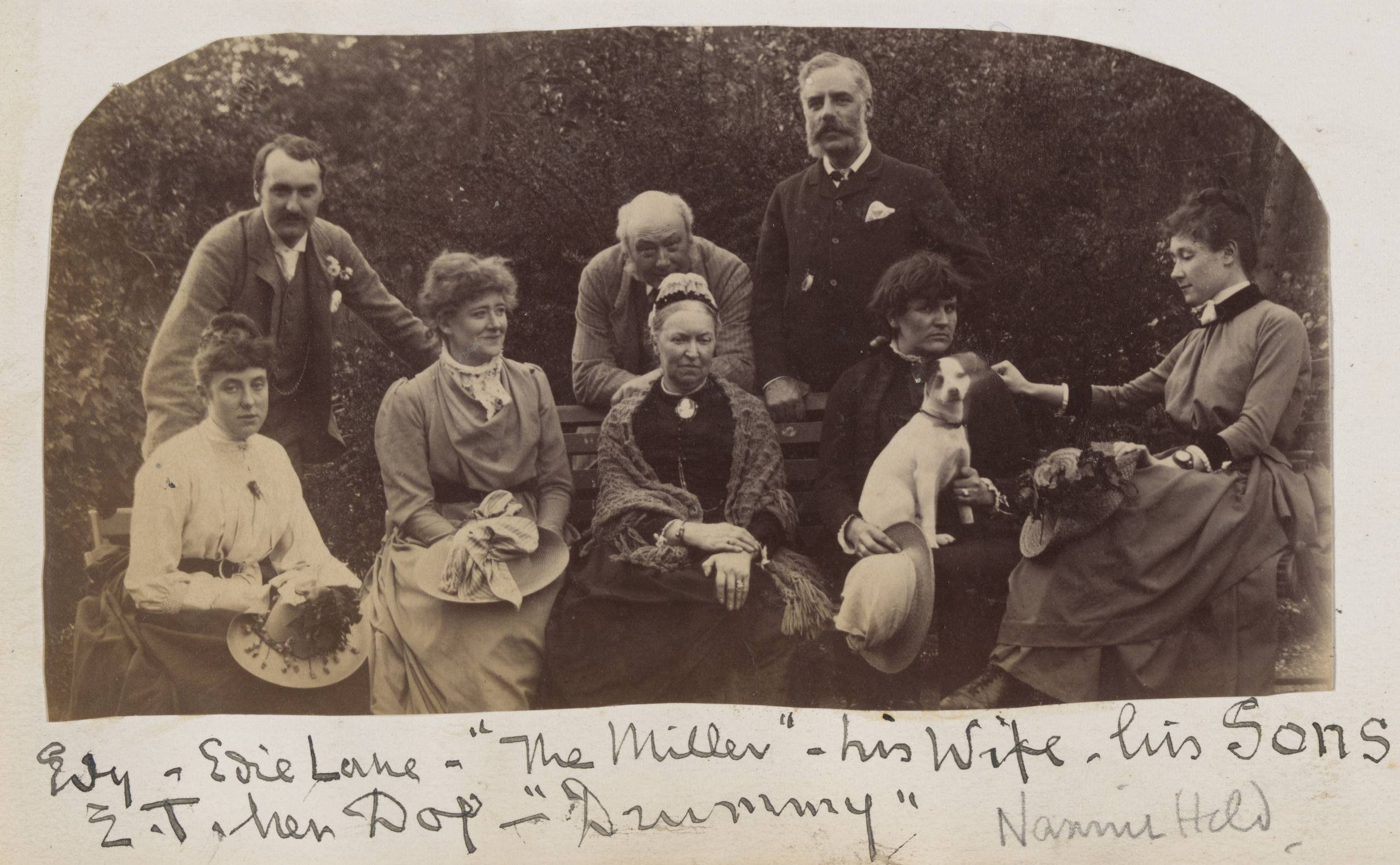 A black and white photograph of a group of five seated women and three standing men. On the lap of the woman fourth from the left is a small white dog with dark ears. The photo is labeled in flowing handwriting.