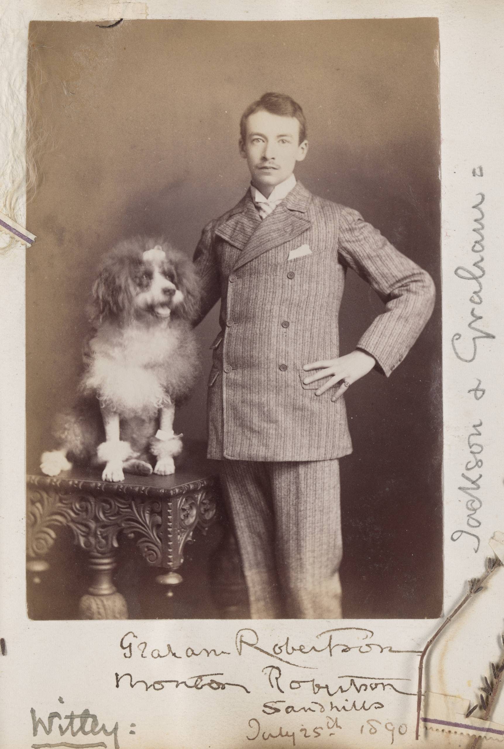 A sepia-toned portrait photograph of a mustachioed man in a suit standing with arm around a well-groomed dog standing on a table.