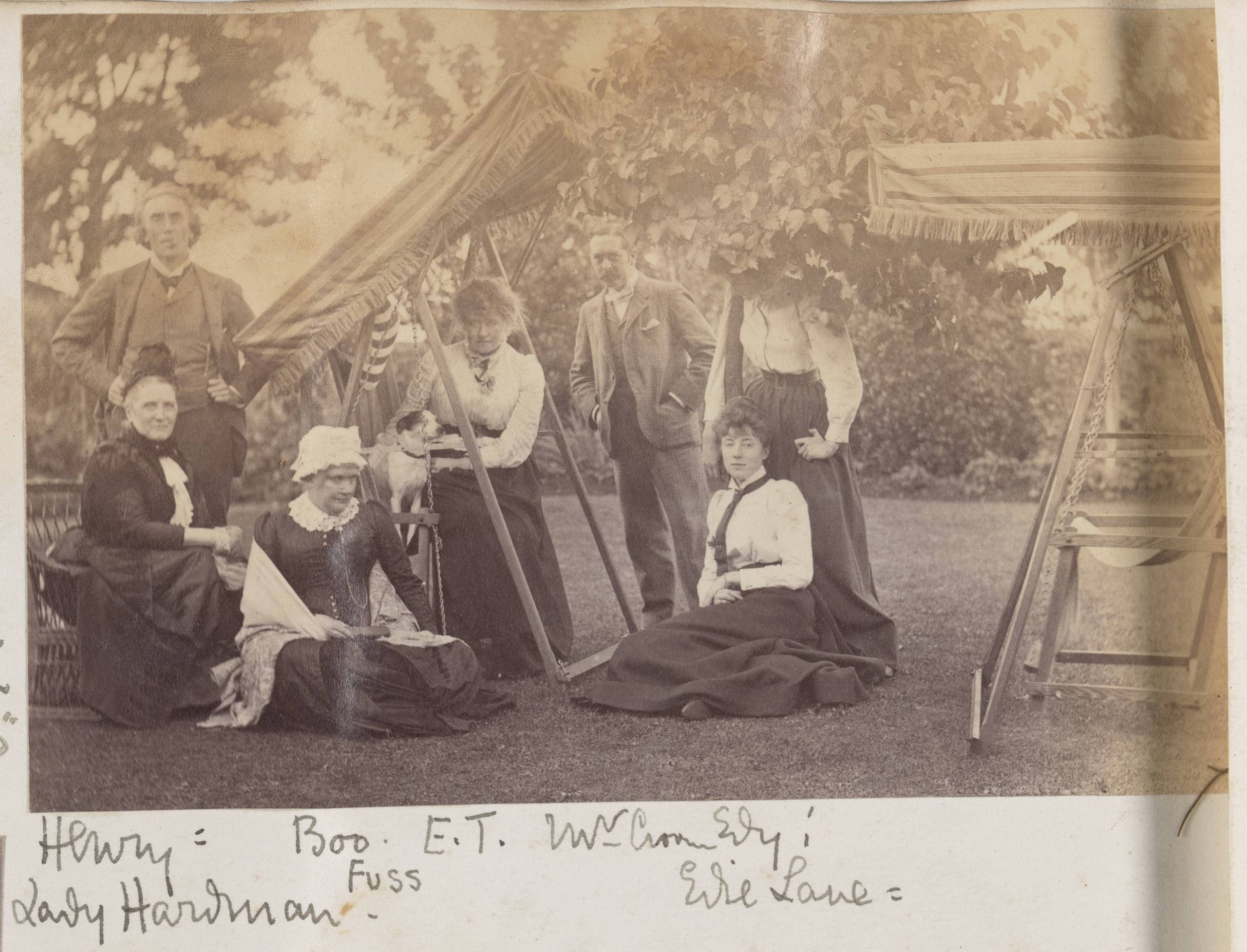A faded sepia-toned photograph of a group of people in a garden seated and standing around garden furniture. A small white dog with dark ears is standing on a chair next to a seated woman. The photo is captioned in flowing handwriting.