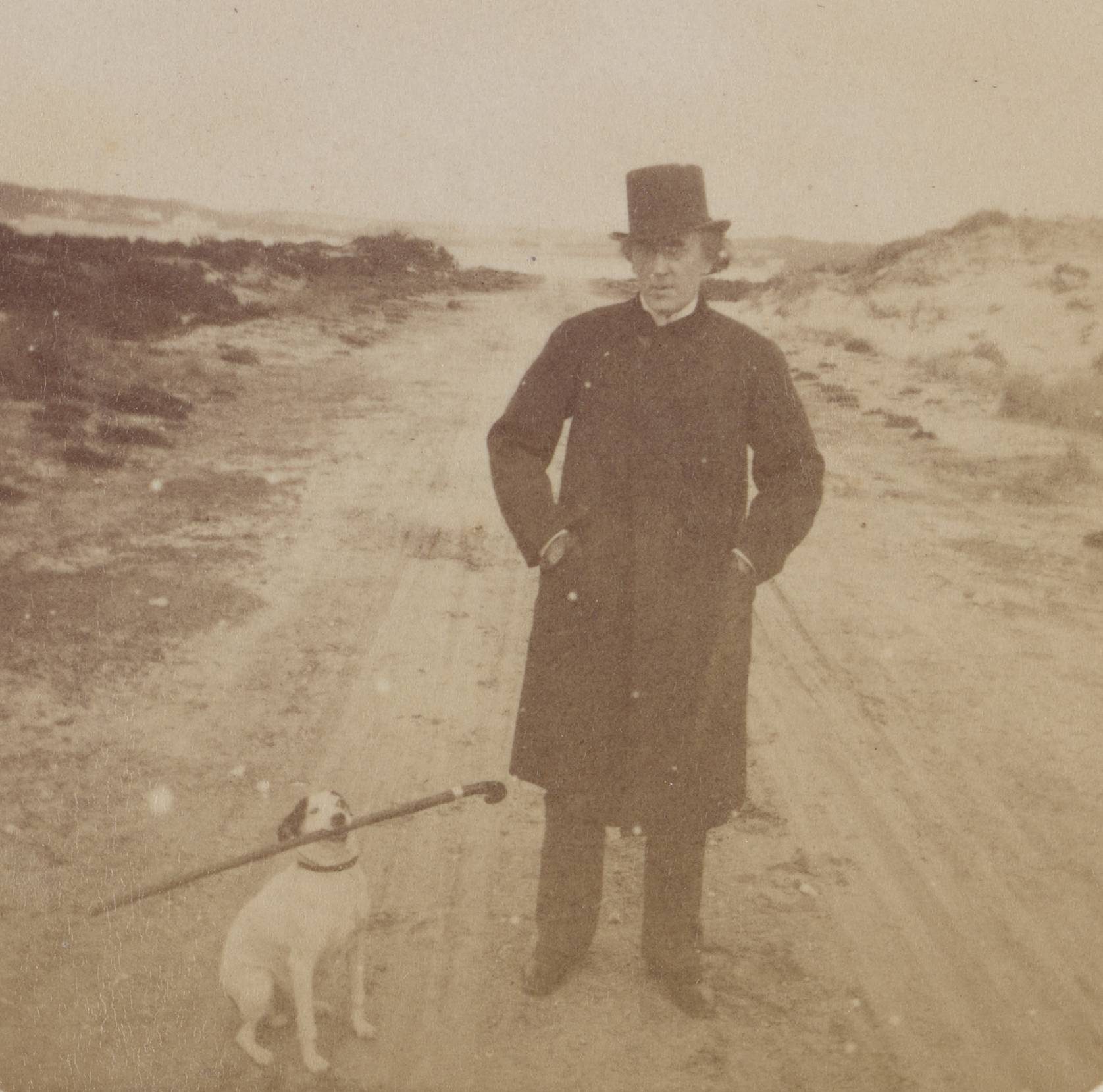 A round faded sepia-toned photograph showing a man standing in the middle of the road. Beside him a small white dog with dark ears holds a cane in its mouth.