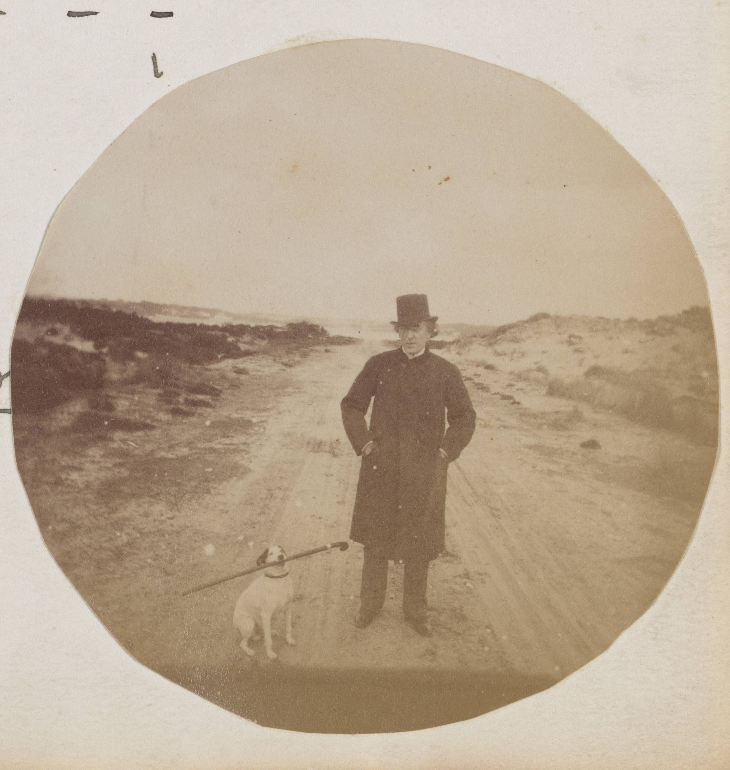 A round faded sepia-toned photograph showing a man standing in the middle of the road. Beside him a small white dog with dark ears holds a cane in its mouth.