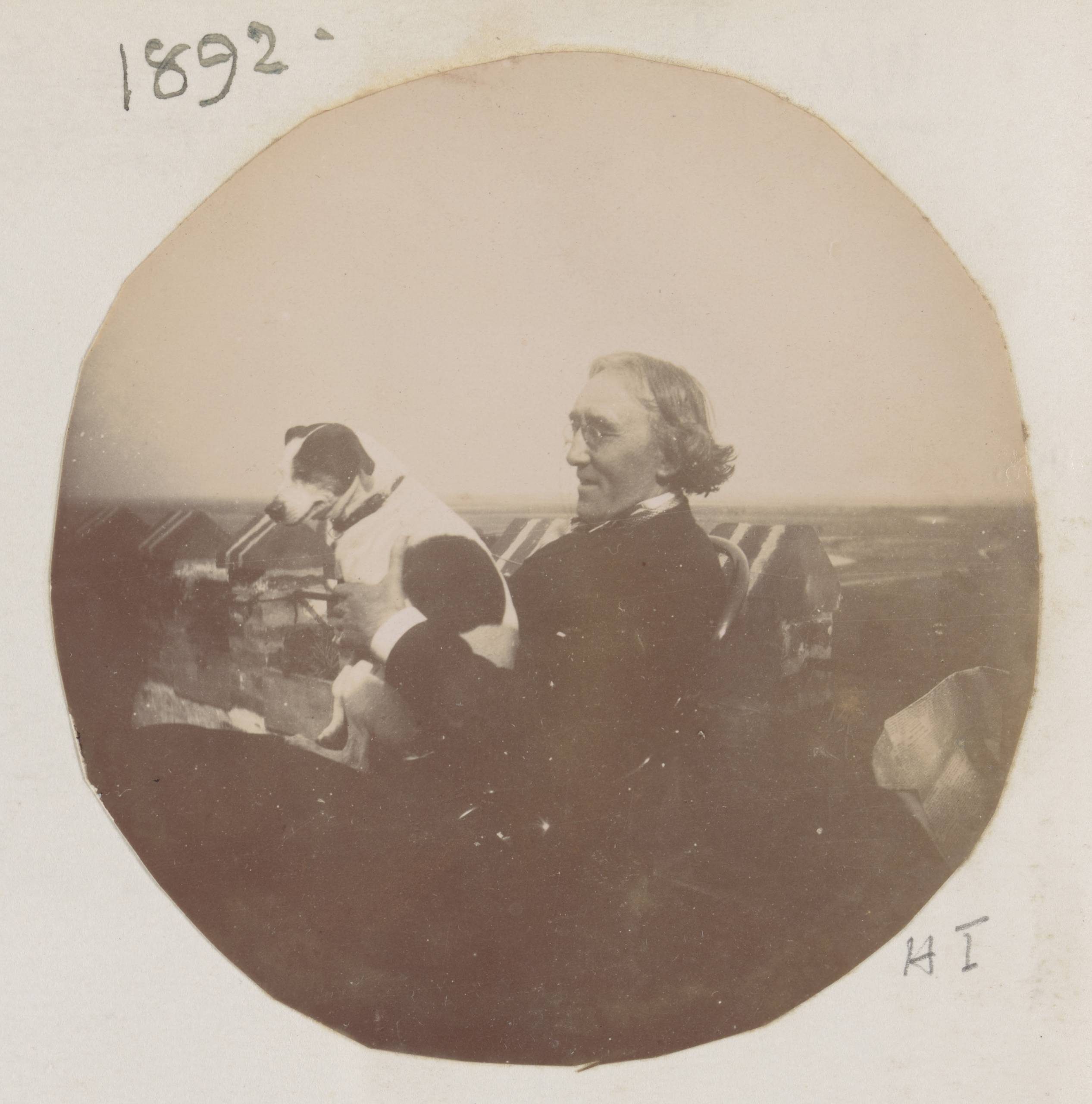 A round sepia-toned photograph of a man seated in a beach chair. On his lap, he holds a small white dog with dark ears and a dark spot on his side.