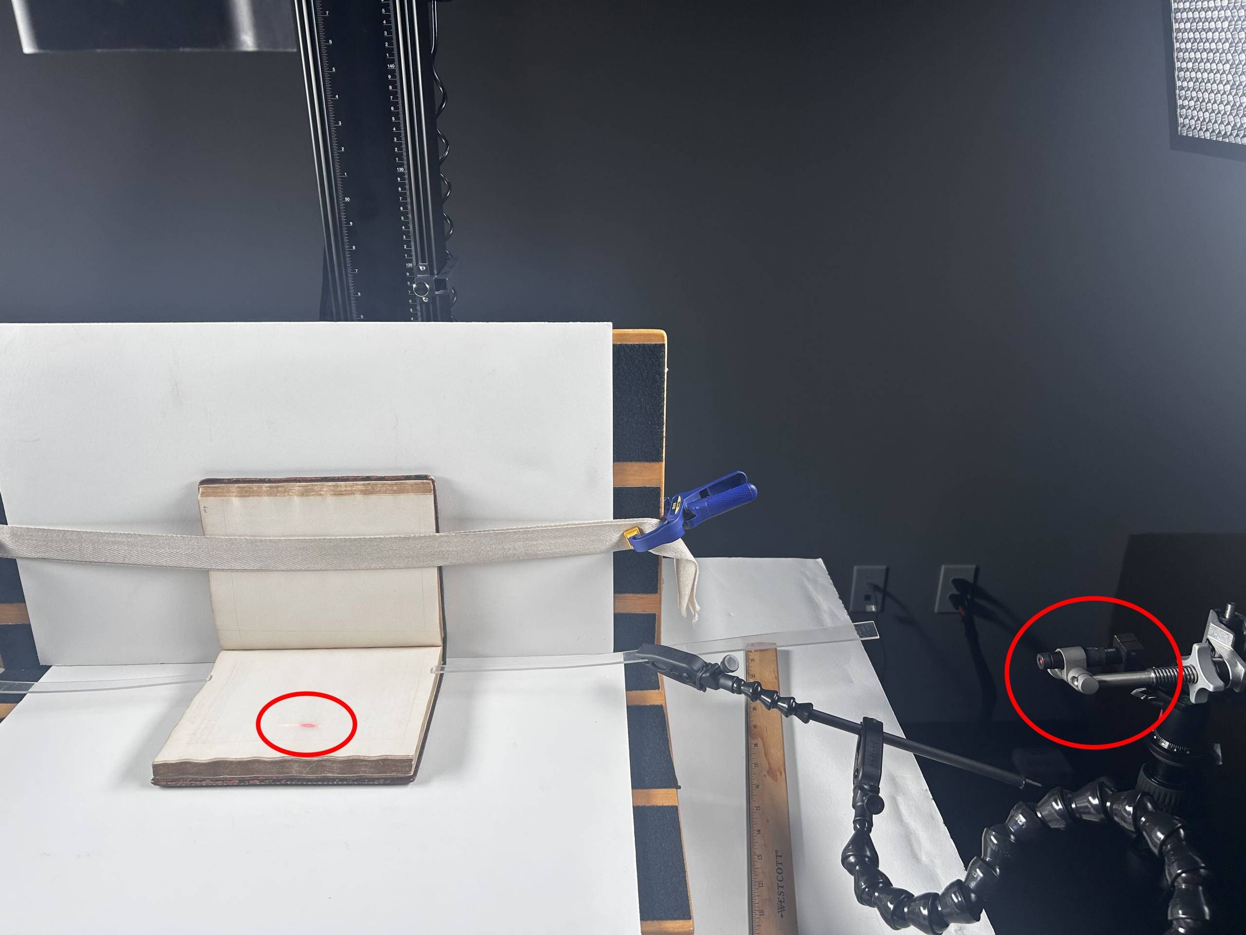 An imaging setup consisting of a table and supports covered in white paper. A book is strapped open on the table. A camera is mounted above the book.